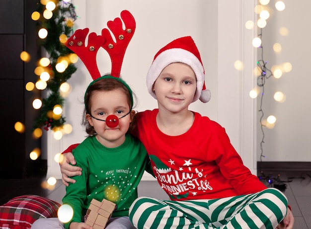 Feliz menina sorridente e menino no chapéu de Papai Noel e máscara de veado, sentado no cobertor de pijama de Natal com caixa de presente.
