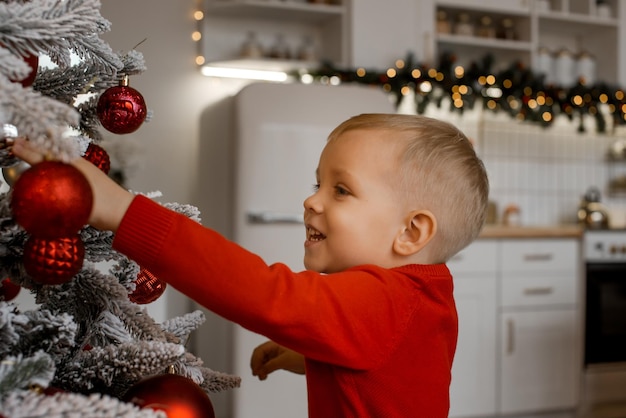 Feliz menino animado no suéter vermelho de Natal está decorando a árvore de Natal no fundo da cozinha desfocada