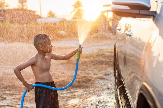 Feliz menino asiático jogando água da mangueira e spray para lavar o carro no exterior em tempo de manhã