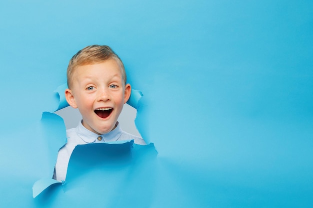 Feliz menino bonitinho está se divertindo brincando na parede de fundo azul sobe por um buraco no papel