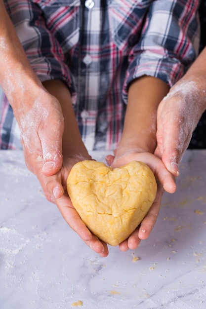 Feliz menino bonito segurando nas mãos um pedaço de massa caseira em forma de coração. Tempo para a família na cozinha aconchegante. Atividade de outono em casa. Treinamento de ajuda e lição de casa para crianças