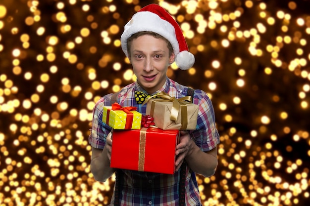Feliz menino caucasiano está segurando uma pilha de presentes. Rapaz bonito com chapéu de Papai Noel. Luzes brilhantes no fundo.