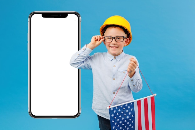 Feliz menino caucasiano no capacete de segurança amarelo segurando e acenando a bandeira americana com rosto sorridente perto do enorme telefone celular