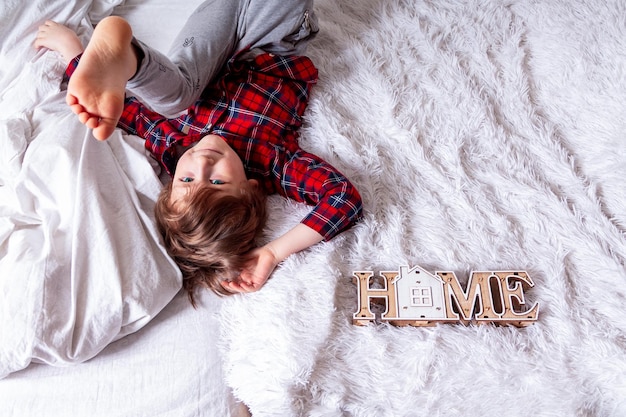 Feliz menino encaracolado com carinha sorridente, deitado na cama branca em casa e pernas para cima, fique em casa