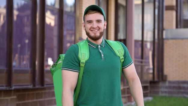 Foto feliz mensajero joven en uniforme verde con una gran bolsa térmica o mochila entregar comida de