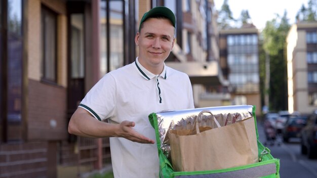 Feliz mensajero joven en uniforme verde con una gran bolsa térmica o mochila entregar comida de