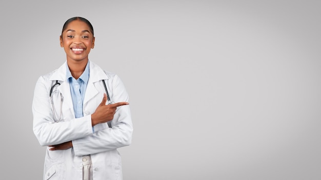 Feliz milenaria hermosa mujer negra médico apunta con el dedo al espacio vacío aislado en el fondo gris