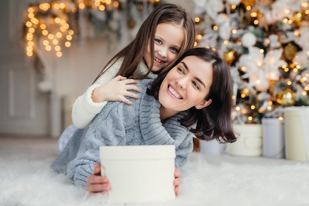 Feliz modelo femenino con cabello corto y oscuro y su adorada niña pequeña se divierten juntos celebran el intercambio de regalos de Navidad tienen expresiones sonrientes tienen alegría Familia amistosa con regalos