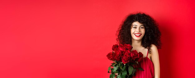 Feliz modelo feminino elegante com buquê de rosas vermelhas sorrindo e olhando alegre para o parafuso prisioneiro da câmera