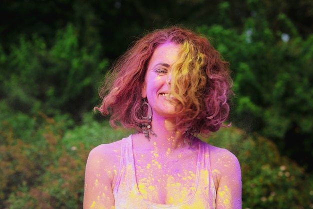 Feliz modelo pelirrojo vistiendo camiseta blanca posando cubierto con pintura colorida Holi seca en el parque