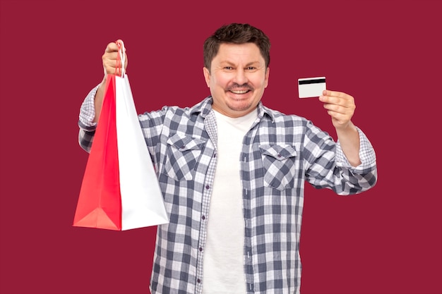 Feliz moderno homem de meia idade em t-shitr branco e camisa quadriculada em pé e segurando sacolas de compras e cartão de crédito com sorriso, olhando para a câmera. estúdio interno filmado em fundo vermelho isolado