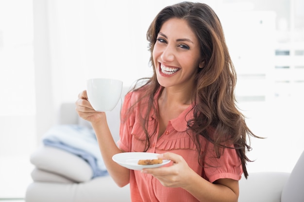 Foto feliz morena sentada en su sofá sosteniendo la taza y el platillo