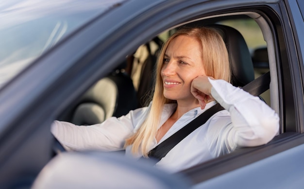 Feliz motorista do sexo feminino com cabelo loiro sentado dentro do carro moderno e olhando direto na estrada mulher madura na camisa azul formal, gostando de dirigir auto sozinha Foto Premium foto