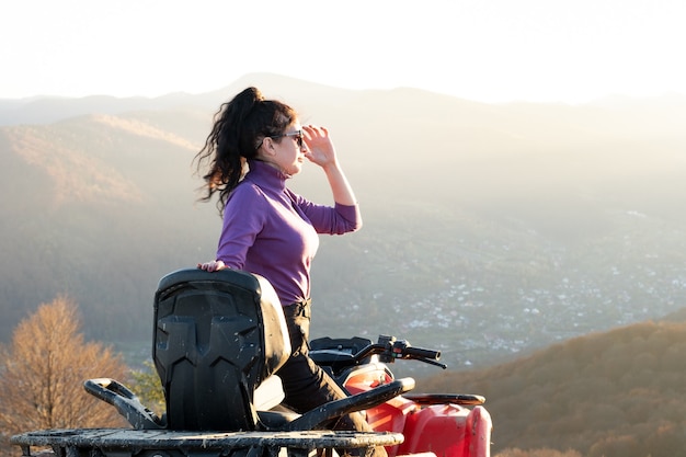 Feliz motorista feminina ativa desfrutando de extrema equitação em moto quadriciclo atv nas montanhas de outono ao pôr do sol.