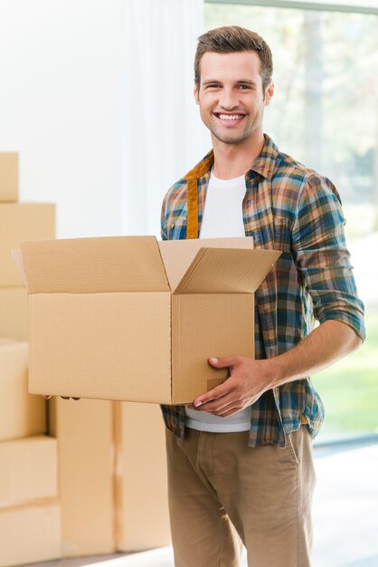 Feliz mudanza a una nueva casa. Apuesto joven sosteniendo una caja de cartón y sonriendo a la cámara mientras otras cajas de cartón ponen en segundo plano.