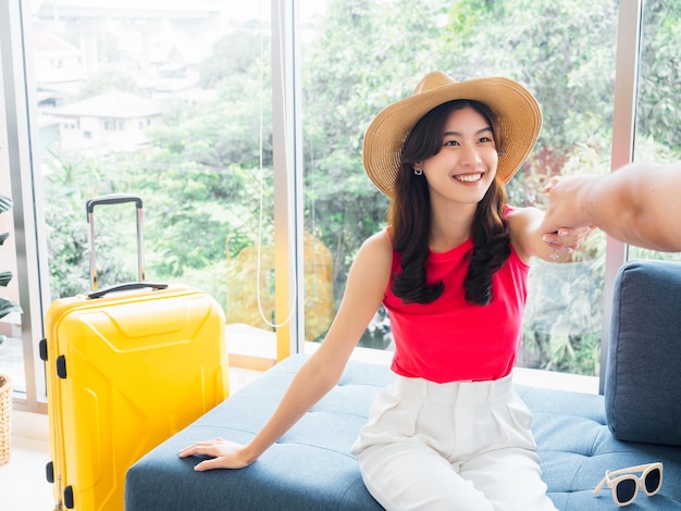 Feliz muito jovem mulher asiática na camisa casual rosa com rosto sorridente senta-se no sofá perto de uma mala amarela apanhada pela mão de alguém para ir em uma viagem closeup sorridente jovem mulher indo viajar