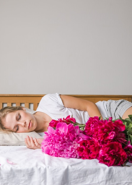 Feliz mujer acostada en la cama en pijama, disfrutando de flores de tulipán ramo de peonía