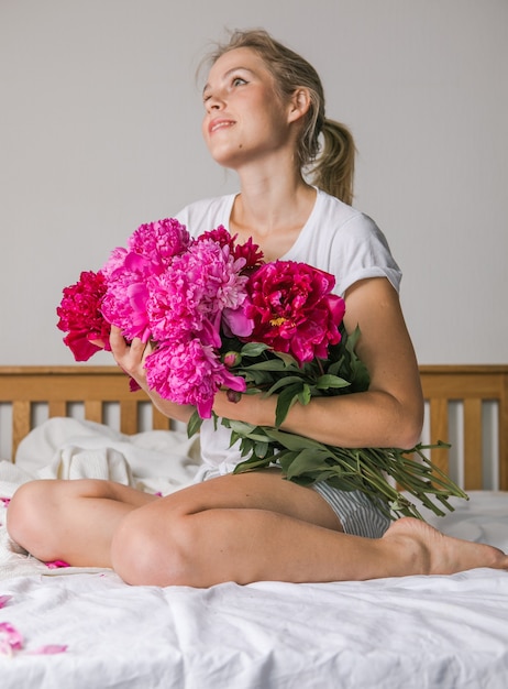 Feliz mujer acostada en la cama en pijama, disfrutando de flores de tulipán ramo de peonía