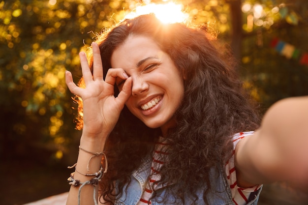 Foto feliz mujer adolescente, a través del signo de ok mientras camina al aire libre y toma una foto selfie