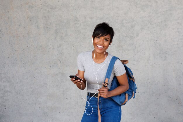 Feliz mujer africana con bolsa de escuchar música en el móvil contra la pared gris