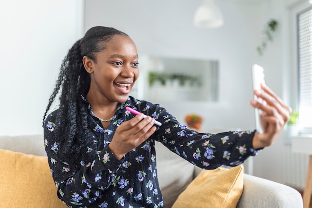 Feliz mujer africana embarazada está mostrando su prueba de embarazo y tomando selfie haciendo videollamadas. Mujer feliz tomando una foto de la prueba de embarazo con el teléfono móvil y publicando una imagen en las redes sociales.