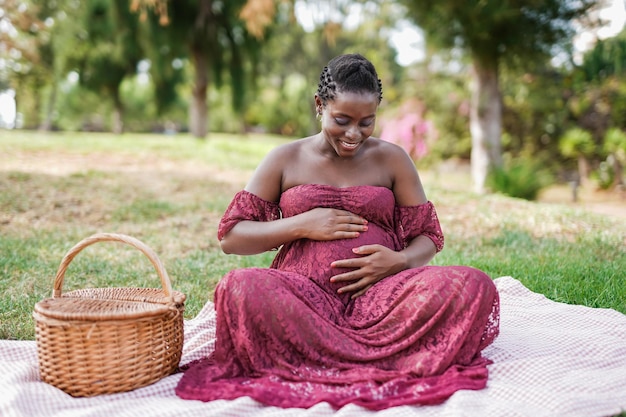 Feliz mujer africana embarazada sentada en el parque de la ciudad y tocándose el vientre