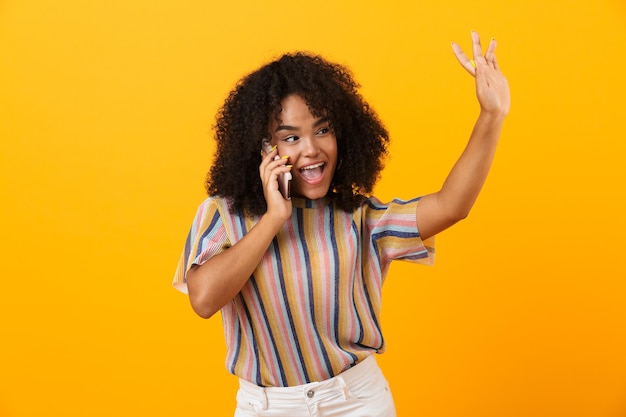 Feliz mujer africana emocional posando aislada sobre espacio amarillo hablando por teléfono móvil.