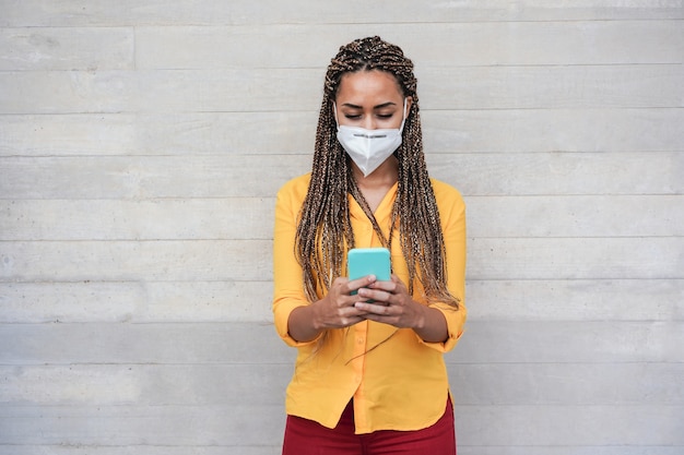 Feliz mujer africana con mascarilla protectora mediante teléfono móvil al aire libre - Centrarse en la cara