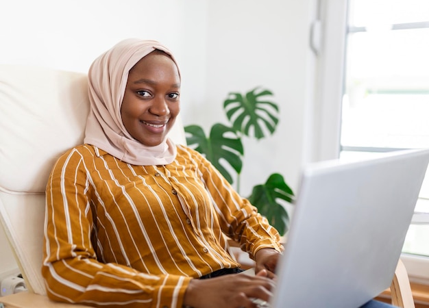 Feliz mujer africana musulmana bonita usando una computadora portátil sentada en un sofá acogedor Hermosa joven musulmana está usando una computadora portátil y sonriendo mientras está sentada en el sofá en casa