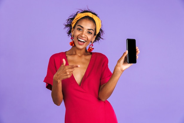 Feliz mujer africana en vestido mostrando la pantalla del smartphone en blanco y apuntando en él