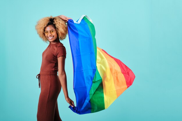 Feliz mujer afro sosteniendo una bandera de orgullo lgbt mientras está de pie sobre un fondo aislado. Concepto de comunidad LGBT.