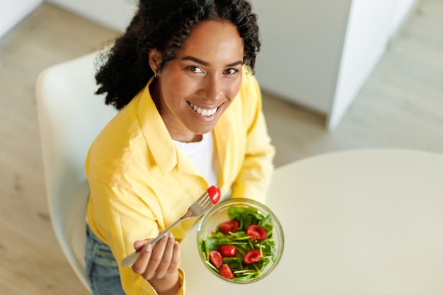 Feliz mujer afroamericana comiendo ensalada con verduras orgánicas y sonriendo a la cámara sentada en la mesa sobre la vista