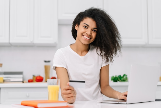 Feliz mujer afroamericana de compras en línea, ordenar comida, de pie en la cocina. Retrato de muchacha hermosa hipster usando laptop, con tarjeta de crédito, haciendo pagos, trabajando desde casa