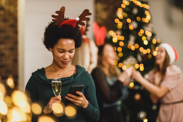 Feliz mujer afroamericana enviando un mensaje de texto en el teléfono inteligente en la fiesta de Año Nuevo en un ambiente festivo.