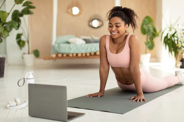 Feliz mujer afroamericana estirándose hacia atrás y viendo tutoriales en línea en una computadora portátil haciendo ejercicio en el dormitorio en una alfombra de yoga