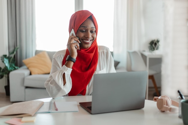 Feliz mujer afroamericana en hijab trabajando en una computadora portátil en la oficina en casa y hablando por teléfono inteligente con el cliente