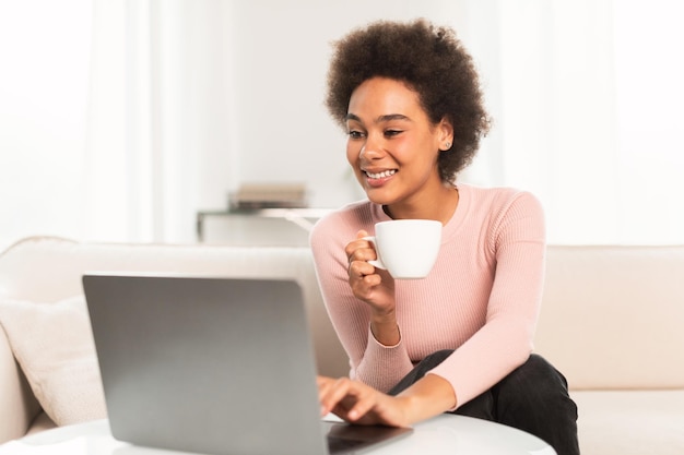 Feliz mujer afroamericana milenaria disfruta de una taza de café y escribe en la computadora portátil en la luminosa sala de estar