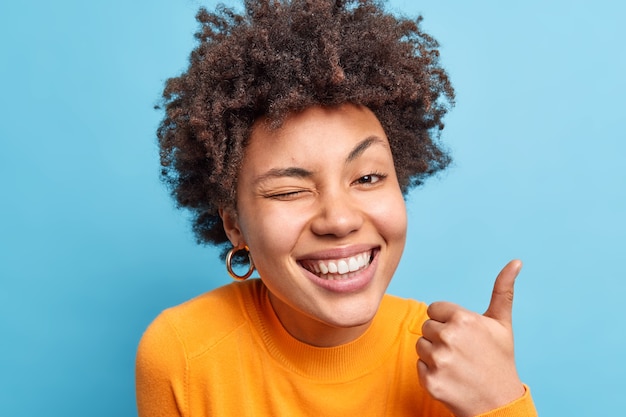 Feliz mujer afroamericana positiva sonríe ampliamente hace un buen gesto con el pulgar hacia arriba dice que sus guiños excelentes ojos sonríen ampliamente usa un jersey naranja aprueba algo aislado en la pared azul