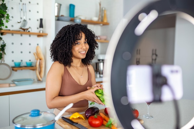 Feliz mujer afroamericana sonriendo y demostrando verduras maduras mientras graba un video para cocinar vlog en la cocina en casa