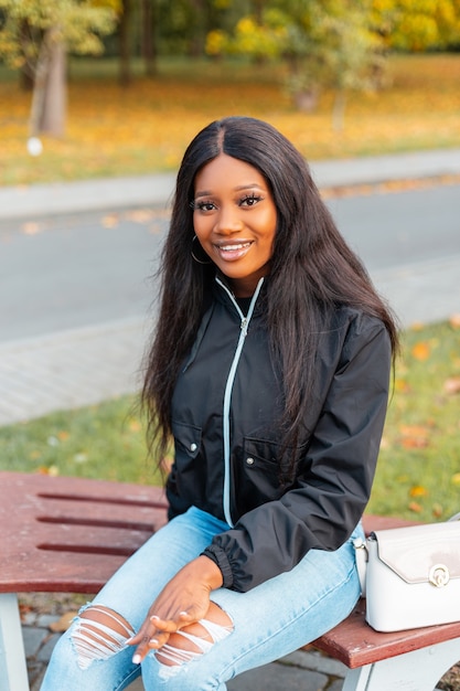 Feliz mujer afroamericana con sonrisa en ropa casual de moda con chaqueta, jeans y bolso sentado en un banco al aire libre