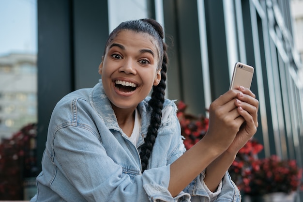 Feliz mujer afroamericana mediante teléfono móvil, mirando a la cámara