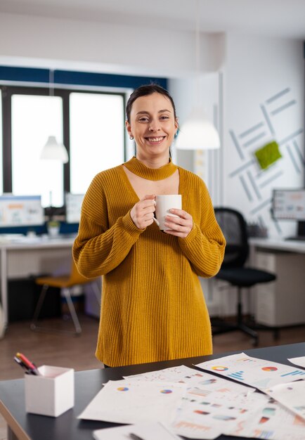 Foto feliz mujer alegre confiada mirando a la cámara en la puesta en marcha de la oficina de la empresa sosteniendo una taza de café. emprendedor ejecutivo, líder gerente permanente trabajando en proyectos.