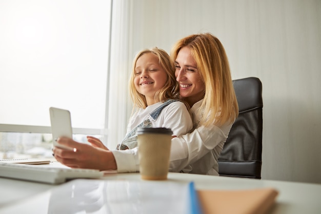 Feliz mujer alegre con niña sentada en la oficina mientras usa el teléfono inteligente moderno