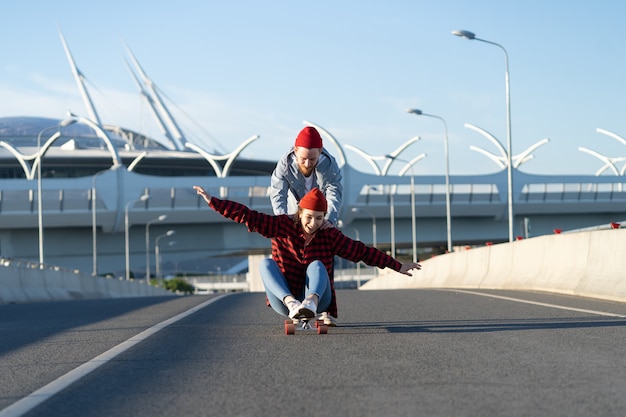 Foto feliz mujer alegre sentarse en longboard mientras el hombre la empuja hacia atrás en el concepto de libertad de las calles de la ciudad