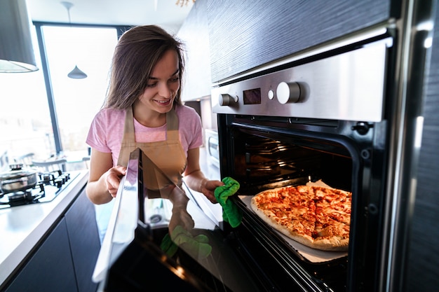 Feliz mujer ama de casa en delantal cocinando pizza casera para la cena con horno en casa