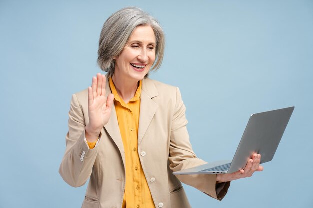 Feliz mujer anciana de cabello gris agitando la mano a la computadora portátil trabajando en línea