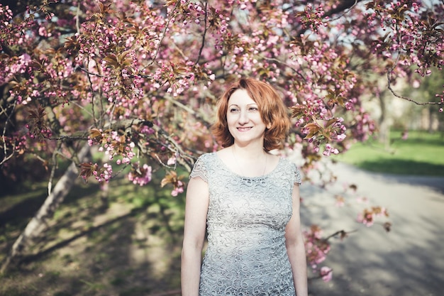 Feliz mujer armenia de mediana edad con un vestido elegante bajo el floreciente árbol de sakura Sonriendo mirando a un lado