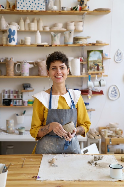 Feliz mujer artista de cerámica que trabaja por cuenta propia en un trabajo de estudio creativo con arcilla cruda que da forma a una taza hecha a mano