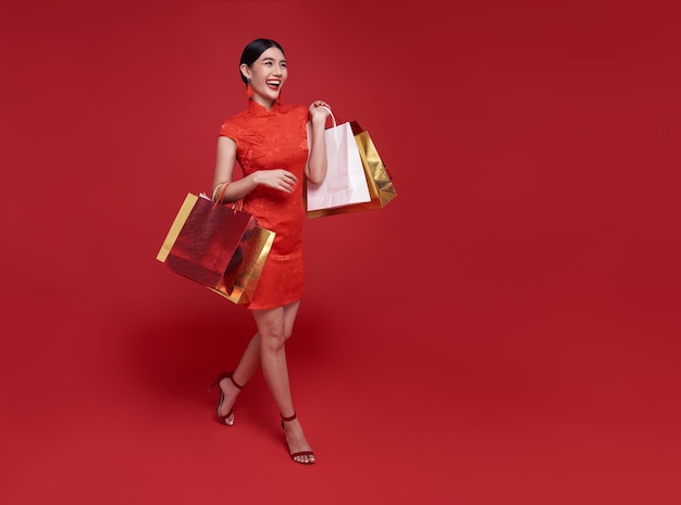 Feliz mujer asiática adicta a las compras con un vestido tradicional cheongsam qipao sosteniendo una bolsa de compras aislada de fondo rojo. Feliz Año Nuevo Chino