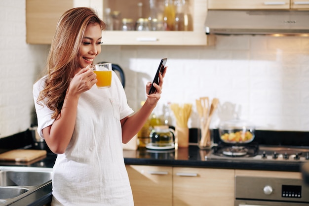 Feliz mujer asiática bastante joven de pie en la cocina, bebiendo una taza de jugo de naranja y leyendo noticias y mensajes de texto en las redes sociales